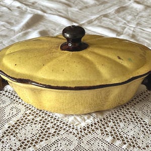 May include: A yellow ceramic casserole dish with a lid, featuring a dark brown knob and rim. The dish has a scalloped design and small handles. It is set on a white lace tablecloth.