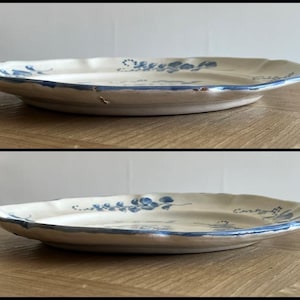 May include: A round ceramic plate with a cream-colored base and a scalloped edge. The plate features hand-painted blue floral motifs and a thin blue line along the rim. The surface shows visible signs of wear and minor chipping along the edge.