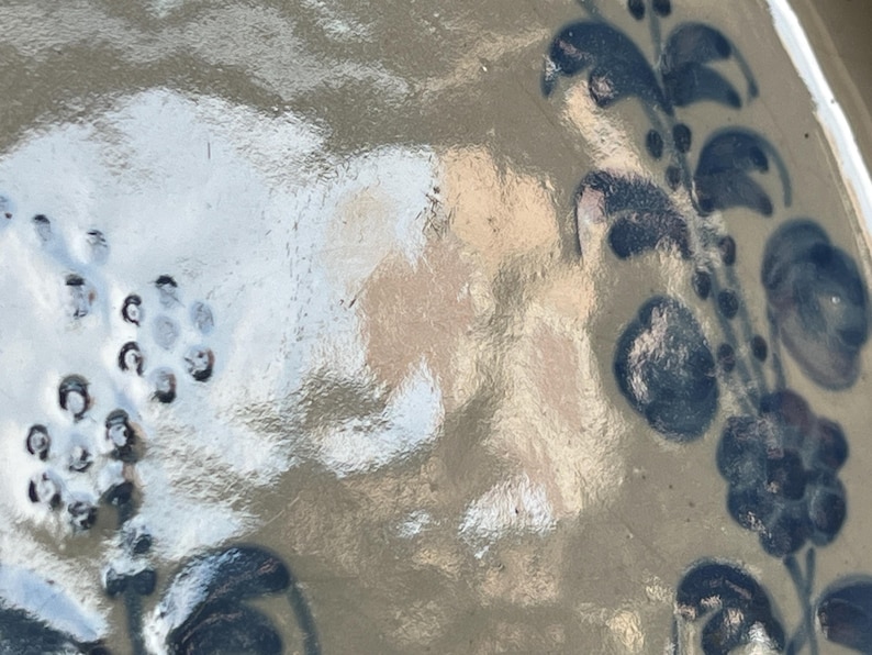 May include: A glazed ceramic surface with a light tan base color featuring hand-painted dark blue floral motifs. The design includes clusters of small circular dots and stylized leaf patterns arranged in a vertical vine-like formation on the right.