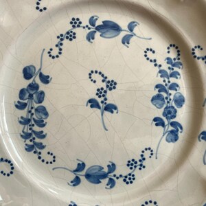 May include: A round ceramic plate with a cream-colored base and a visible crackle glaze finish. The plate features hand-painted blue floral motifs, including delicate vines, leaves, and small clusters of berries arranged in a circular pattern.