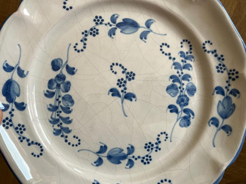 May include: A round ceramic plate with a cream-colored base and a visible crackle glaze finish. The plate features hand-painted blue floral motifs, including delicate vines, leaves, and small clusters of berries arranged in a circular pattern.