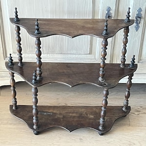 Può includere: Una scaffalatura a tre livelli in legno marrone scuro con bordi smerlati su ogni ripiano. I ripiani sono sostenuti da pilastri torniti a forma di fuso con terminali decorativi che si estendono verso l'alto dal ripiano superiore.
