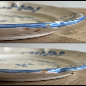 May include: A cream-colored ceramic plate featuring a blue floral pattern and a blue painted rim. The plate has a scalloped edge and visible surface crazing. There are two distinct chips along the outer rim where the ceramic is exposed.