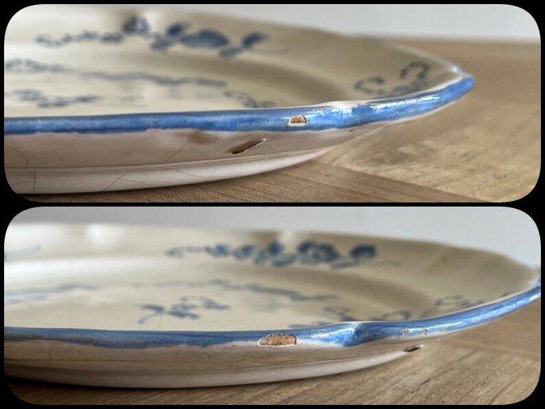May include: A cream-colored ceramic plate featuring a blue floral pattern and a blue painted rim. The plate has a scalloped edge and visible surface crazing. There are two distinct chips along the outer rim where the ceramic is exposed.