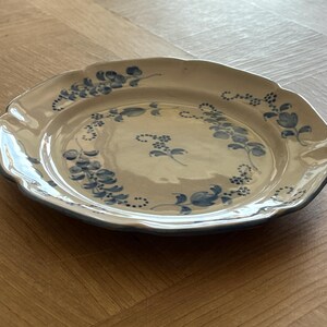 May include: A round ceramic plate with a scalloped edge, featuring a cream-colored base and a hand-painted blue floral pattern. The design includes delicate blue vines and small blossoms arranged in a circular motif around the center.