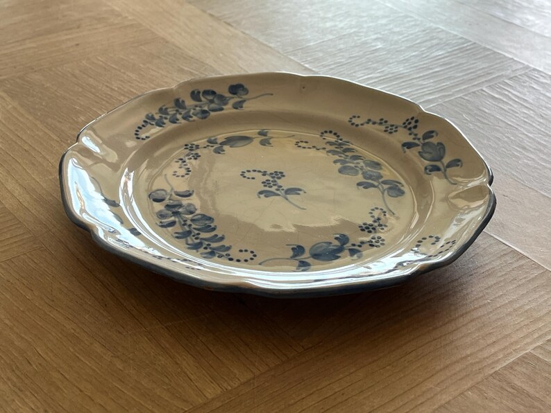 May include: A round ceramic plate with a scalloped edge, featuring a cream-colored base and a hand-painted blue floral pattern. The design includes delicate blue vines and small blossoms arranged in a circular motif around the center.