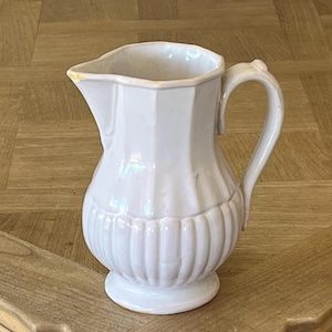 May include: White ceramic pitcher with a handle and a wide spout. The pitcher has a ribbed design around the base and a faceted body. The handle is curved, and the pitcher sits on a small, round base. The pitcher is likely for serving beverages.