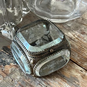 May include: A square glass jewellery box featuring bevelled edges and intricate floral etchings on the lid and sides. The glass panels are held together by an ornate, dark metallic frame with a textured, scalloped border and a small central clasp.