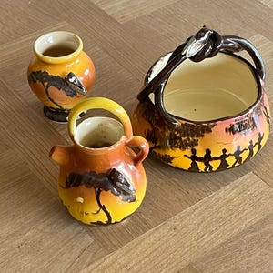 Op de afbeelding: Drie keramische vazen met een zonsondergangontwerp. De vazen zijn in tinten geel, oranje en bruin. Eén vaas is een kleine, ronde pot. Een andere is een kleine kan met een handvat. De derde is een mand met een handvat.