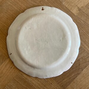 May include: The underside of a round, off-white ceramic plate featuring a scalloped edge and a visible network of fine cracks across the glaze. A small metal hanging loop is embedded into the top rim, allowing the plate to be mounted on a wall.