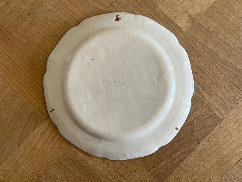 May include: The underside of a round, off-white ceramic plate featuring a scalloped edge and a visible network of fine cracks across the glaze. A small metal hanging loop is embedded into the top rim, allowing the plate to be mounted on a wall.