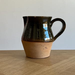 May include: A handmade ceramic pitcher with a dark green glazed top and a natural tan base. The pitcher has a curved handle and a spout for pouring. This rustic piece is suitable for serving beverages.