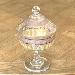 Puede incluir: Un bombonera de cristal transparente con tapa. El plato tiene una base de pedestal y está decorado con un patrón floral rosa y dorado. La tapa está rematada con un pomo de cristal transparente.