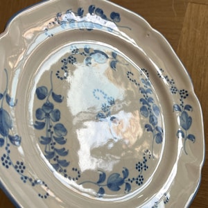 May include: A cream-colored ceramic plate with a scalloped edge and a blue hand-painted floral pattern. The surface features a visible crackle glaze finish, with blue painted vines and small flower clusters decorating the center and rim.