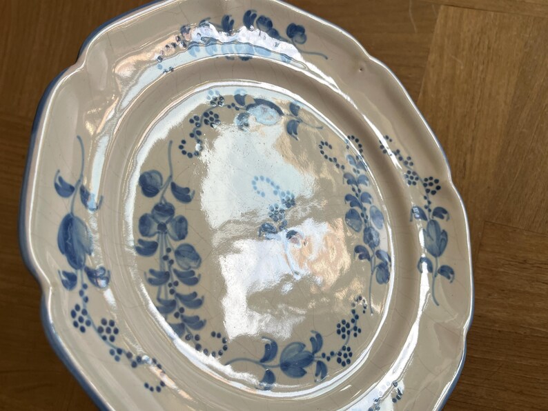 May include: A cream-colored ceramic plate with a scalloped edge and a blue hand-painted floral pattern. The surface features a visible crackle glaze finish, with blue painted vines and small flower clusters decorating the center and rim.