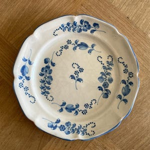 May include: A round ceramic plate with a scalloped edge, featuring a white base with a blue hand-painted floral pattern. The design includes delicate vines and small blossoms arranged in a circular motif, with visible fine crackle glazing.