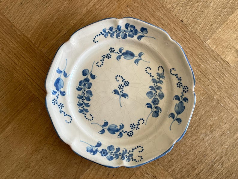May include: A round ceramic plate with a scalloped edge, featuring a white base with a blue hand-painted floral pattern. The design includes delicate vines and small blossoms arranged in a circular motif, with visible fine crackle glazing.