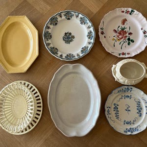 May include: A collection of seven ceramic pieces including a yellow octagonal platter, a white plate with blue floral patterns, a white plate with a pink rose motif, a cream lidded tureen, a cream lattice-rimmed plate, a plain oval platter, and a blue floral plate.