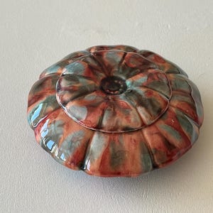 May include: A ceramic trinket box with a lid. The box is round with a floral design and is glazed in shades of red, teal, and brown. The lid has a small, central knob.