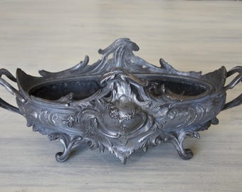May include: A silver-plated, ornate planter with a decorative floral design and two handles. The planter has a scalloped edge and sits on three feet.