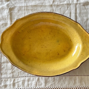 May include: Oval yellow ceramic serving dish with a scalloped edge and a dark brown rim. The dish has a slightly textured surface and sits on a white, patterned tablecloth. A vintage piece for serving food.
