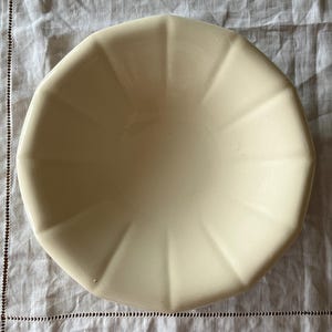 Op de afbeelding: Een crèmekleurige, achthoekige keramische kom met een gegroefd ontwerp. De kom is leeg en staat op een witte, getextureerde tafelkleed. De vorm en kleur van de kom suggereren een vintage of antieke esthetiek.