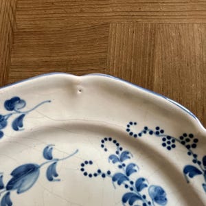 May include: A cream-colored ceramic plate with a scalloped edge and a blue hand-painted floral pattern. The surface features a visible network of fine cracks in the glaze, known as crazing, and a small indentation along the rim.