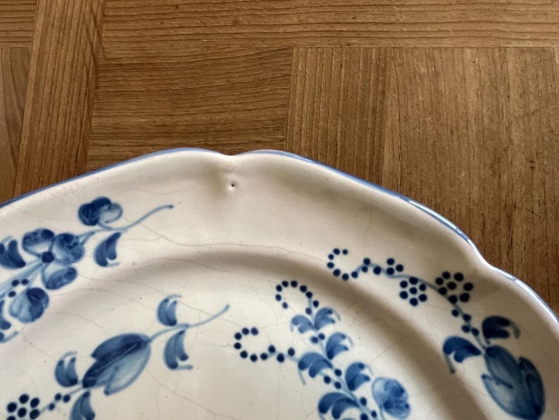 May include: A cream-colored ceramic plate with a scalloped edge and a blue hand-painted floral pattern. The surface features a visible network of fine cracks in the glaze, known as crazing, and a small indentation along the rim.