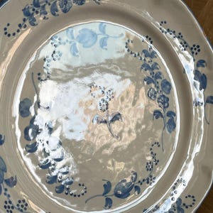 May include: A round ceramic plate with a scalloped edge, featuring a glossy off-white glaze and hand-painted blue floral motifs. The design includes delicate vines, leaves, and small blossoms arranged in a circular pattern around the center.