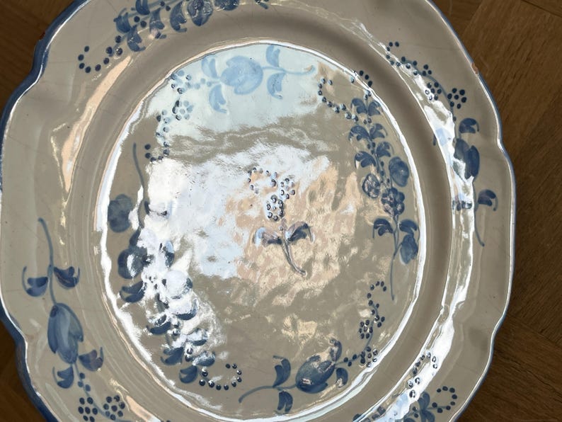 May include: A round ceramic plate with a scalloped edge, featuring a glossy off-white glaze and hand-painted blue floral motifs. The design includes delicate vines, leaves, and small blossoms arranged in a circular pattern around the center.