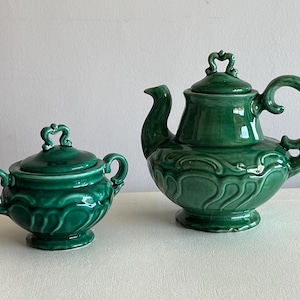 Può includere: Una teiera e una zuccheriera in ceramica verde con dettagli decorativi. La teiera ha un beccuccio curvo e un manico sul lato. La zuccheriera ha un coperchio con un pomo in cima. Entrambi i pezzi hanno un design testurizzato a forma di onda.