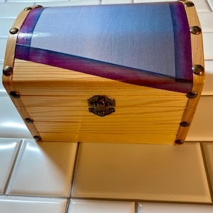May include: A small wooden chest with a hinged lid. The chest is made of light-colored wood and features a decorative design on the lid with shades of blue and purple. The chest is secured with a metal clasp and accented with brass-colored studs.