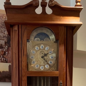 May include: A traditional wooden grandfather clock with a glass door revealing a gold-toned clock face with Roman numerals and a moon phase display. The clock has a brass pendulum and weights.
