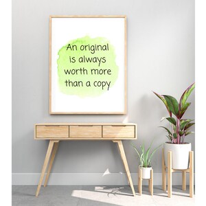 Peut inclure: Une impression encadr&eacute;e avec un fond aquarelle vert et le texte "An original is always worth more than a copy". L'impression est accroch&eacute;e &agrave; un mur blanc au-dessus d'une console en bois avec deux plantes sur des supports en bois.