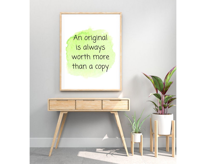 Peut inclure: Une impression encadr&eacute;e avec un fond aquarelle vert et le texte "An original is always worth more than a copy". L'impression est accroch&eacute;e &agrave; un mur blanc au-dessus d'une console en bois avec deux plantes sur des supports en bois.