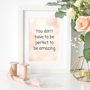 Peut inclure: Impression encadr&eacute;e blanche avec un fond aquarelle et le texte "You don't have to be perfect to be amazing". L'impression est pos&eacute;e sur une surface blanche avec un bougeoir en m&eacute;tal dor&eacute; et deux bobines de ruban rose.