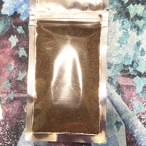 May include: A small, rectangular, silver-colored, sealed pouch filled with dark brown granules. The pouch is transparent, allowing the contents to be visible. The background features a colorful, patterned fabric with shades of blue, purple, and green.