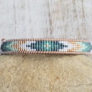 Pode incluir: Um bracelete de contas com um padrão geométrico em tons de verde, branco e dourado. O bracelete é feito com contas pequenas e redondas e tem um suporte de couro.