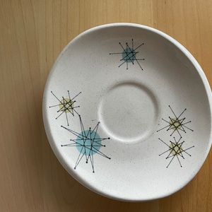 May include: A white saucer with a speckled finish and a black and blue starburst design. The saucer has a center well for a cup.