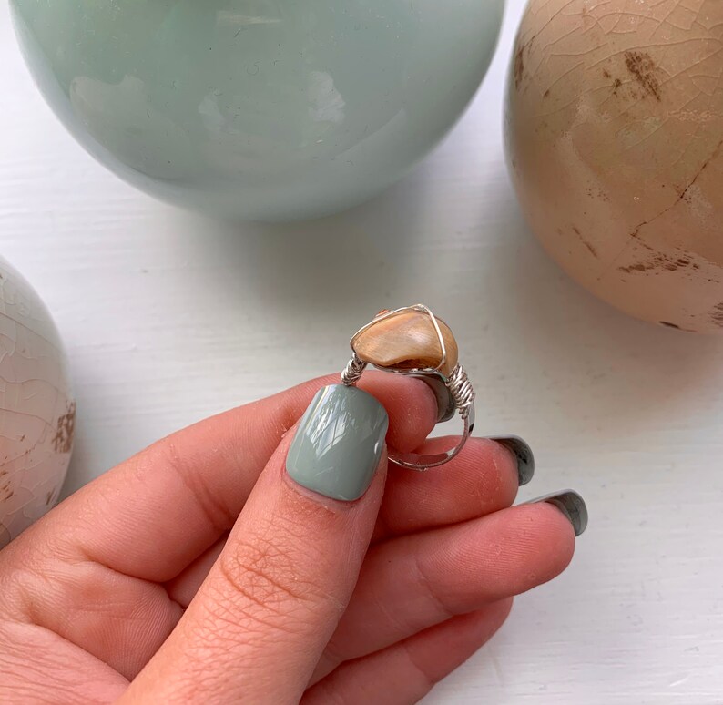 K&ouml;nnte beinhalten: Ein Ring aus silbernem Draht mit einem hellbraunen Stein. Der Ring wird von einer Hand mit hellgr&uuml;nem Nagellack gehalten.