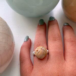 K&ouml;nnte beinhalten: Ein Ring aus silbernem Draht, der mit einer beigefarbenen Muschel als Mittelst&uuml;ck umwickelt ist. Der Ring befindet sich an einem Finger.