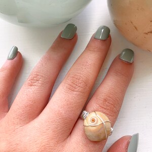 K&ouml;nnte beinhalten: Ein Ring aus einer beigefarbenen Muschel, die mit silbernem Draht umwickelt ist. Der Ring befindet sich an einem Finger mit gr&uuml;nem Nagellack.