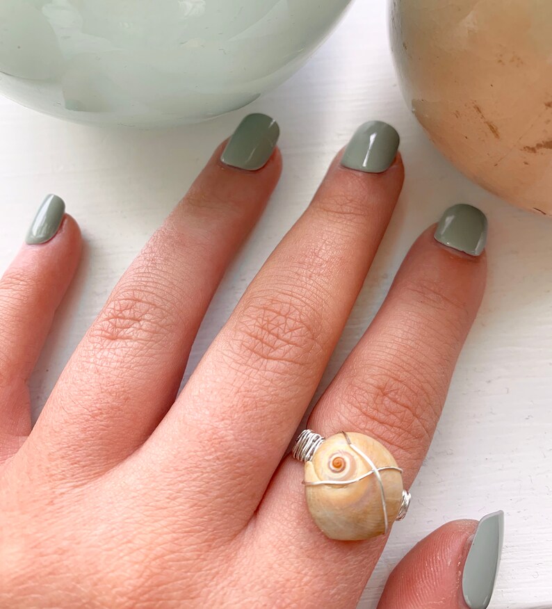 K&ouml;nnte beinhalten: Ein Ring aus einer beigefarbenen Muschel, die mit silbernem Draht umwickelt ist. Der Ring befindet sich an einem Finger mit gr&uuml;nem Nagellack.