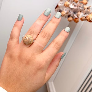 K&ouml;nnte beinhalten: Ein Ring mit einer beigefarbenen Muschel, die mit silbernem Draht umwickelt ist. Der Ring wird an einer Hand mit hellgr&uuml;nem Nagellack getragen.