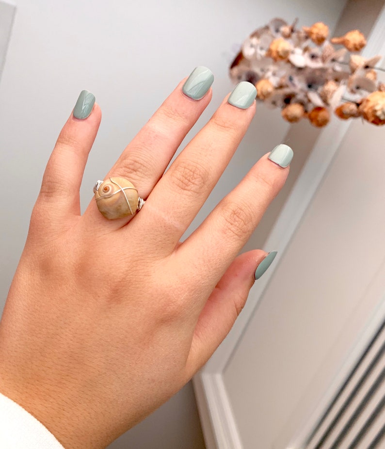 K&ouml;nnte beinhalten: Ein Ring mit einer beigefarbenen Muschel, die mit silbernem Draht umwickelt ist. Der Ring wird an einer Hand mit hellgr&uuml;nem Nagellack getragen.