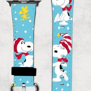 Puede incluir: Una banda de reloj Apple azul y blanca con un patrón de Snoopy con un gorro y una bufanda rojos y blancos. La banda tiene una hebilla plateada.