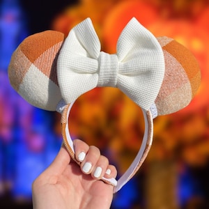 May include: Orange and white plaid Minnie Mouse ears with a large white bow. The ears are on a headband.