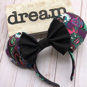 May include: A pair of mouse ears with a large black bow. The ears are covered in a purple fabric with a colorful villain print. The headband is black and the word "dream" is in the background.