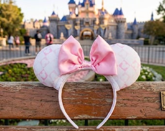 May include: Pink and white Minnie Mouse ear headband with a large pink satin bow. The ears have a white background with a pink diamond pattern and Mickey Mouse head silhouettes. The headband is white and sits on a wooden bench.