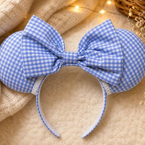 May include: A blue and white gingham patterned headband with large round ears and a matching bow. The headband has a white base with a delicate lace trim. The ears and bow are made of a light blue and white checkered fabric.